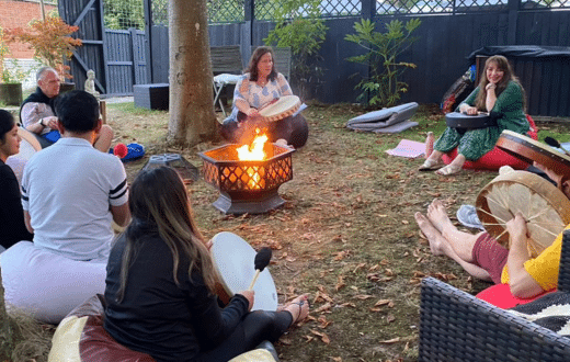 A group of people sit in a circle around a firepit outdoors, playing hand drums and talking, with trees and fencing in the background.
