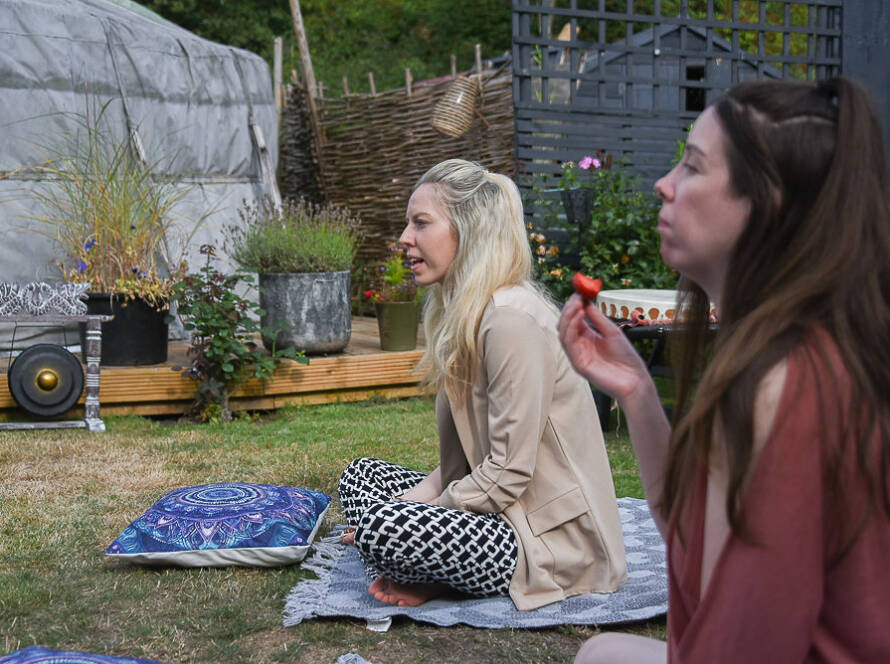 Two women sitting outdoors on a lawn, one eating a strawberry, with cushions, plants, and a gong in the background.