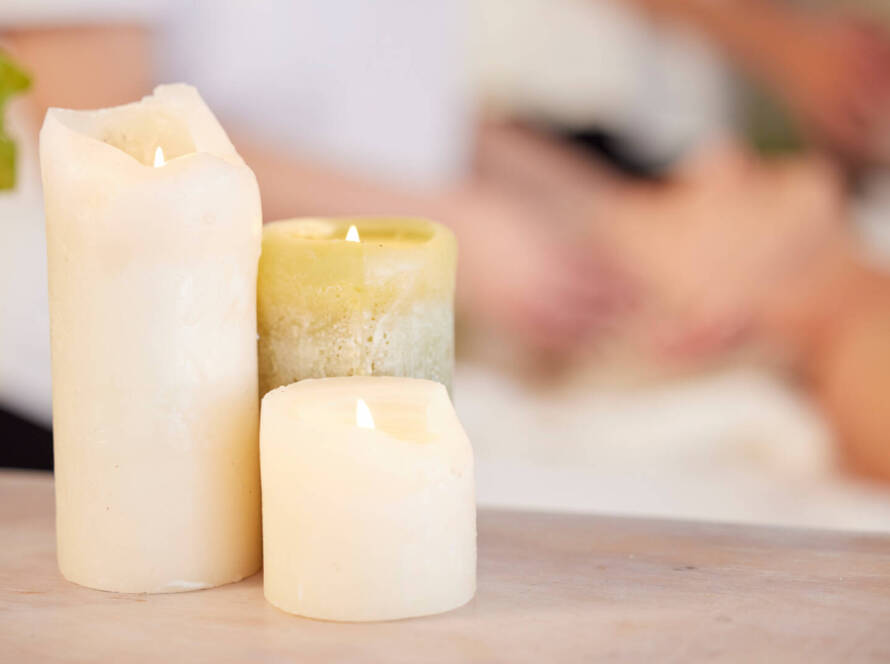 Three lit candles of varying sizes and colours are placed on a table, with a blurred background showing a person receiving a facial treatment.