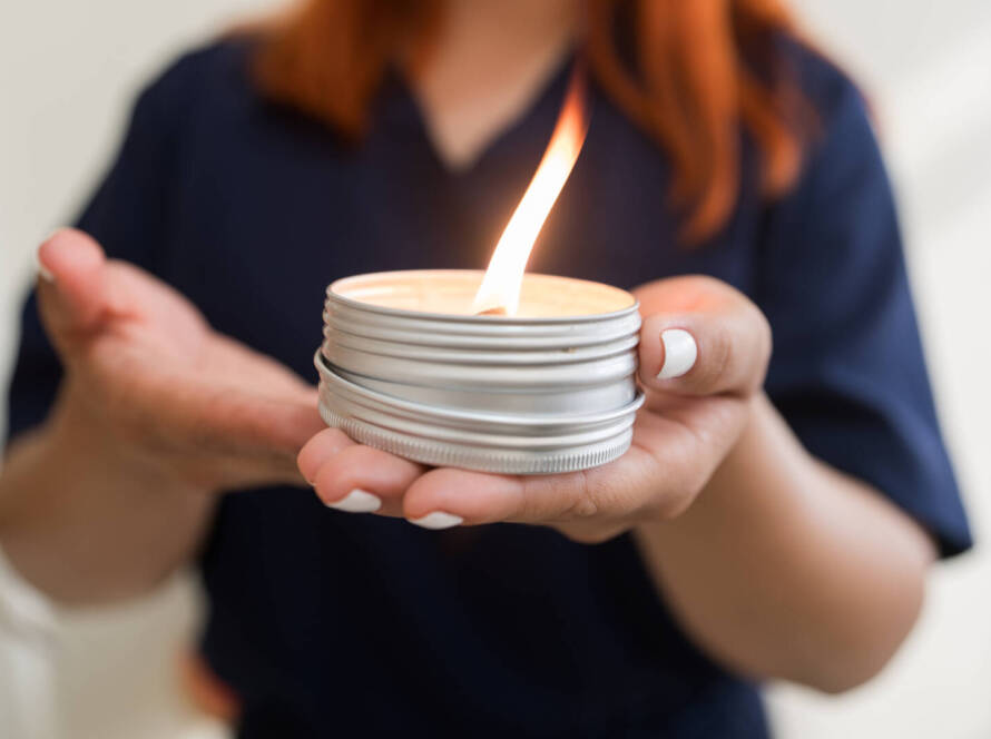 A person with painted nails holds a lit candle in a small metal tin, with flames burning brightly.