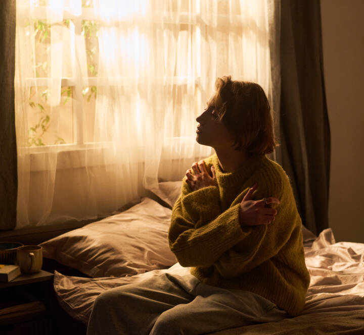 Person in a yellow jumper sits on a bed hugging themselves, looking out a sunlit window with sheer curtains.