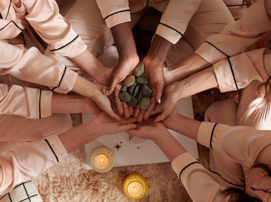 Several people in matching pyjamas form a circle, placing their hands together over a pile of stones, with candles and a rug visible below.
