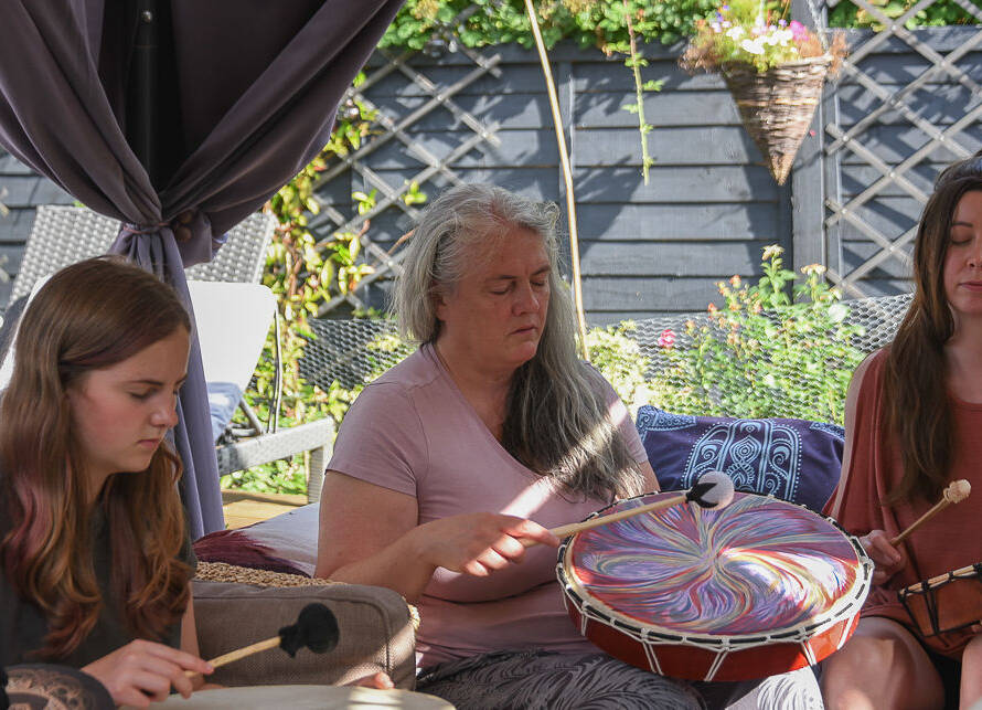 Three women sit outdoors, eyes closed, playing colourful hand drums with mallets. Sunlight filters through, and plants and garden furniture are visible in the background.