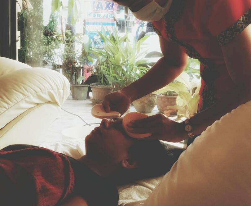 A person lies on a sofa whilst another person wearing a mask applies facial pads to their face in a spa-like setting.