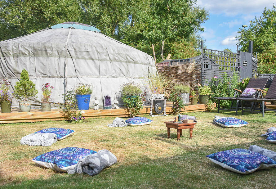 A circular tent is set up in a garden with scattered cushions and blankets on the grass, surrounded by potted plants and wooden outdoor furniture.