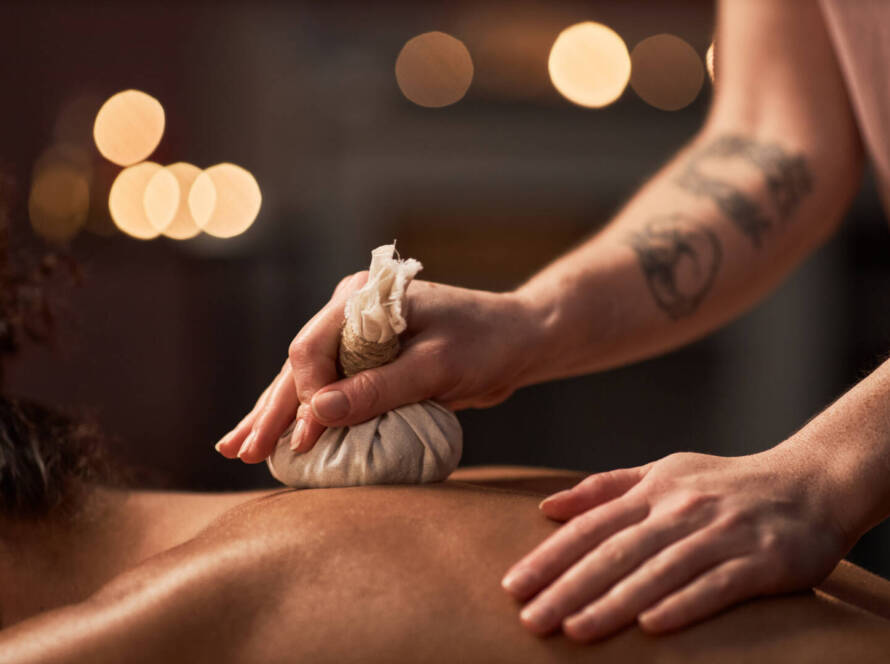 A person receives a massage with a herbal compress on their back, administered by someone with a tattooed arm in a softly lit setting.