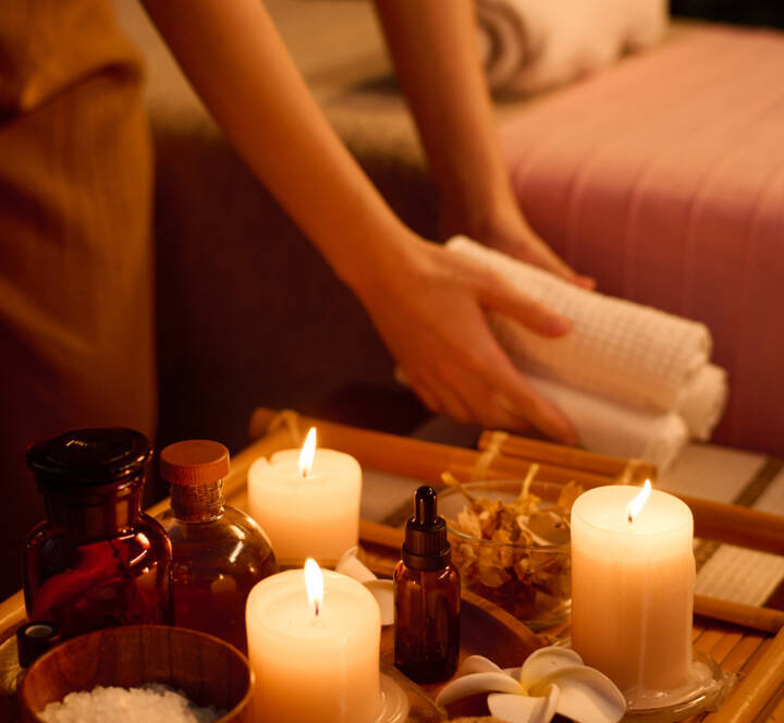 A person arranging towels near a tray with lit candles, glass bottles, a bowl of salt, a dropper bottle, and a scourer in a spa setting.
